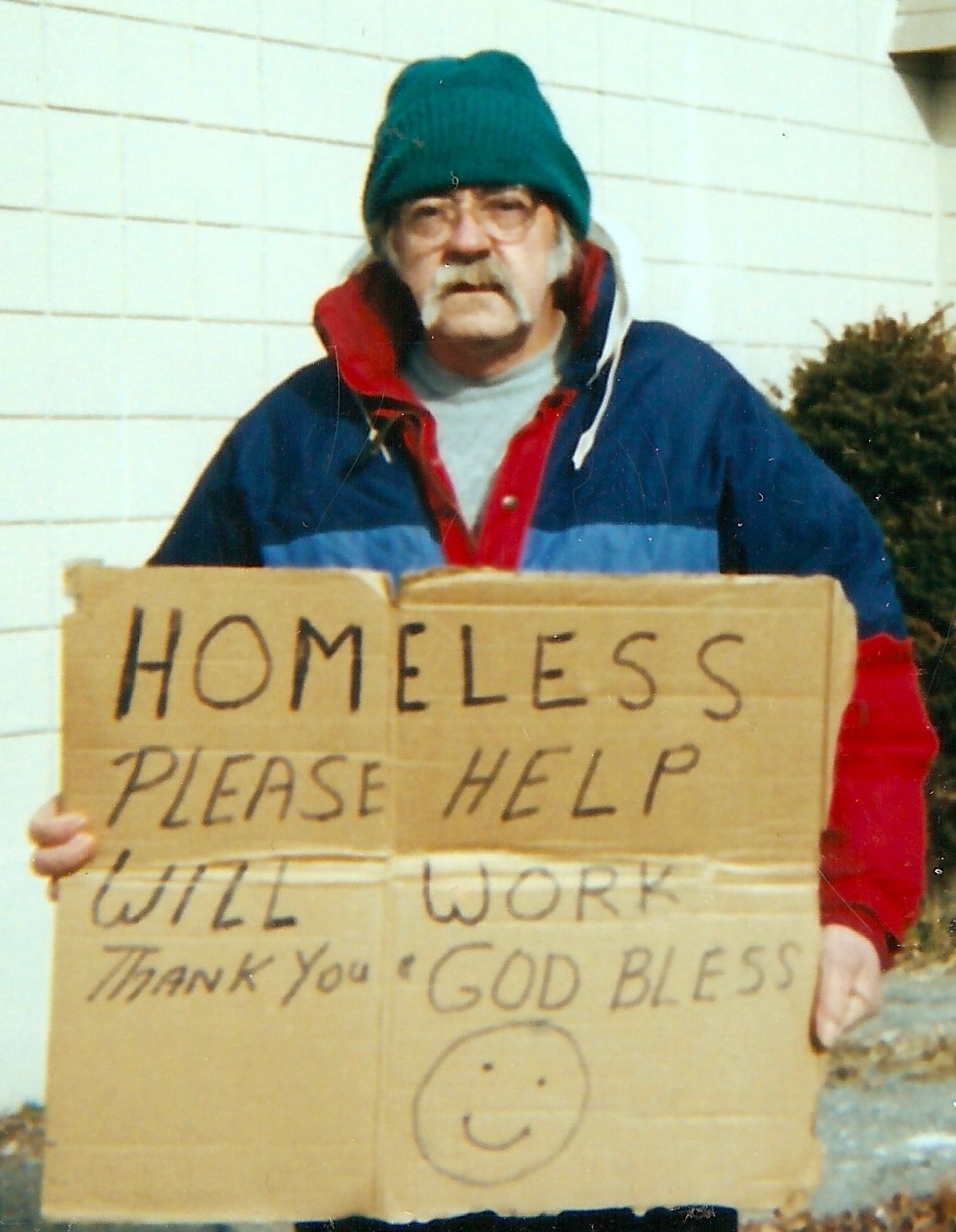 An older white man with a white mustache and glasses. He's wearing a green hat and blue and red jacket. He's holding a sign that reads, 'Homeless, please help. Will work. Thank you & God Bless'